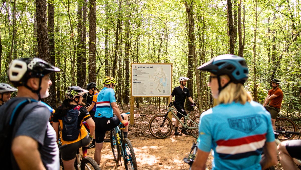 Group of bicyclists standing on bikes around park sign in woods