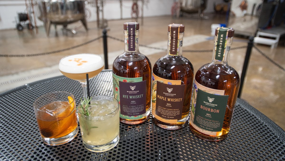 Cocktails and bottles of spirits displayed on table with distilling equipment in background